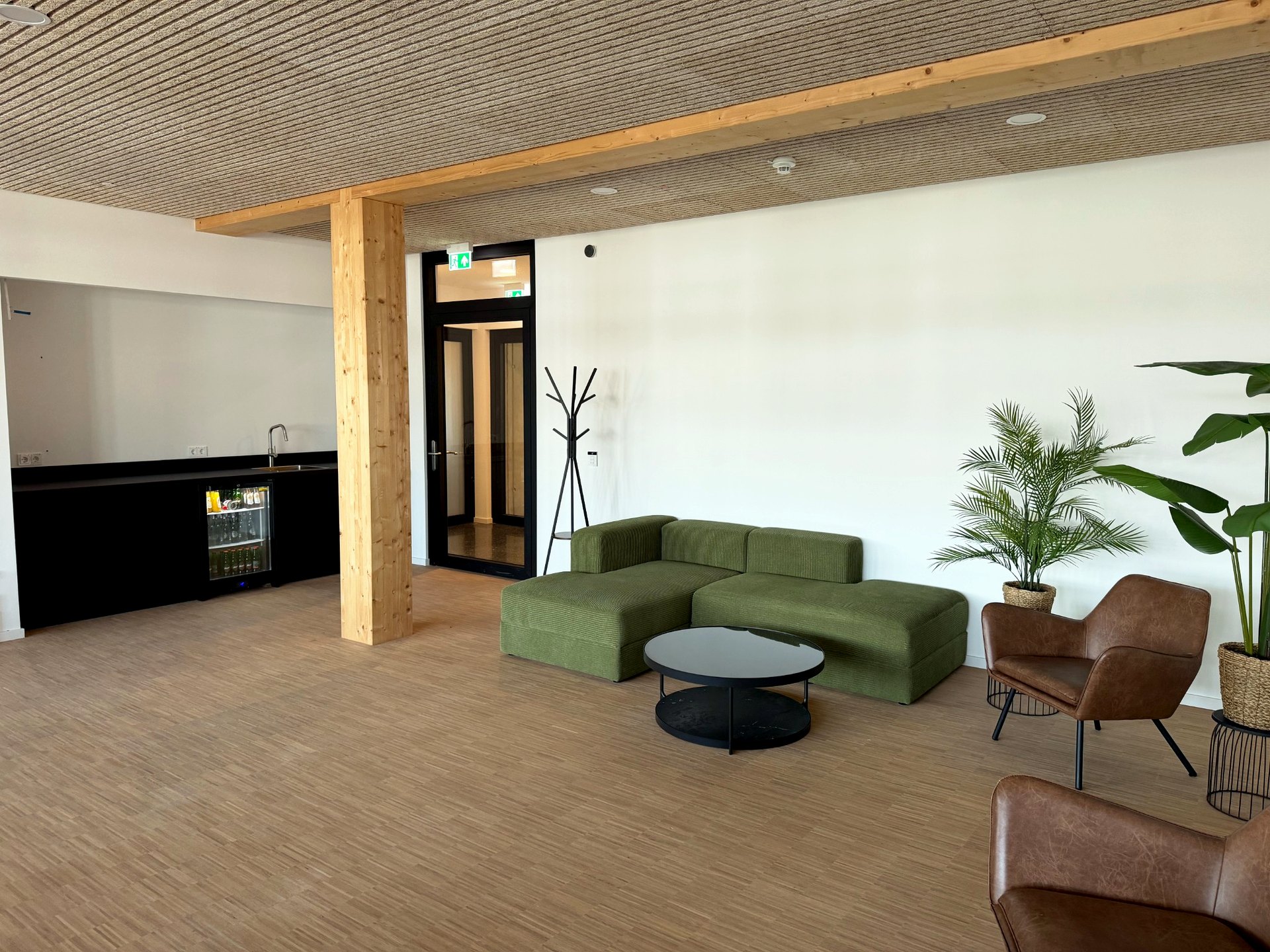 Lounge-Bereich der THINKBOX mit grünem Cord-Sofa, Ledersesseln, Pflanzen und Küchenzeile vor Holzdecke